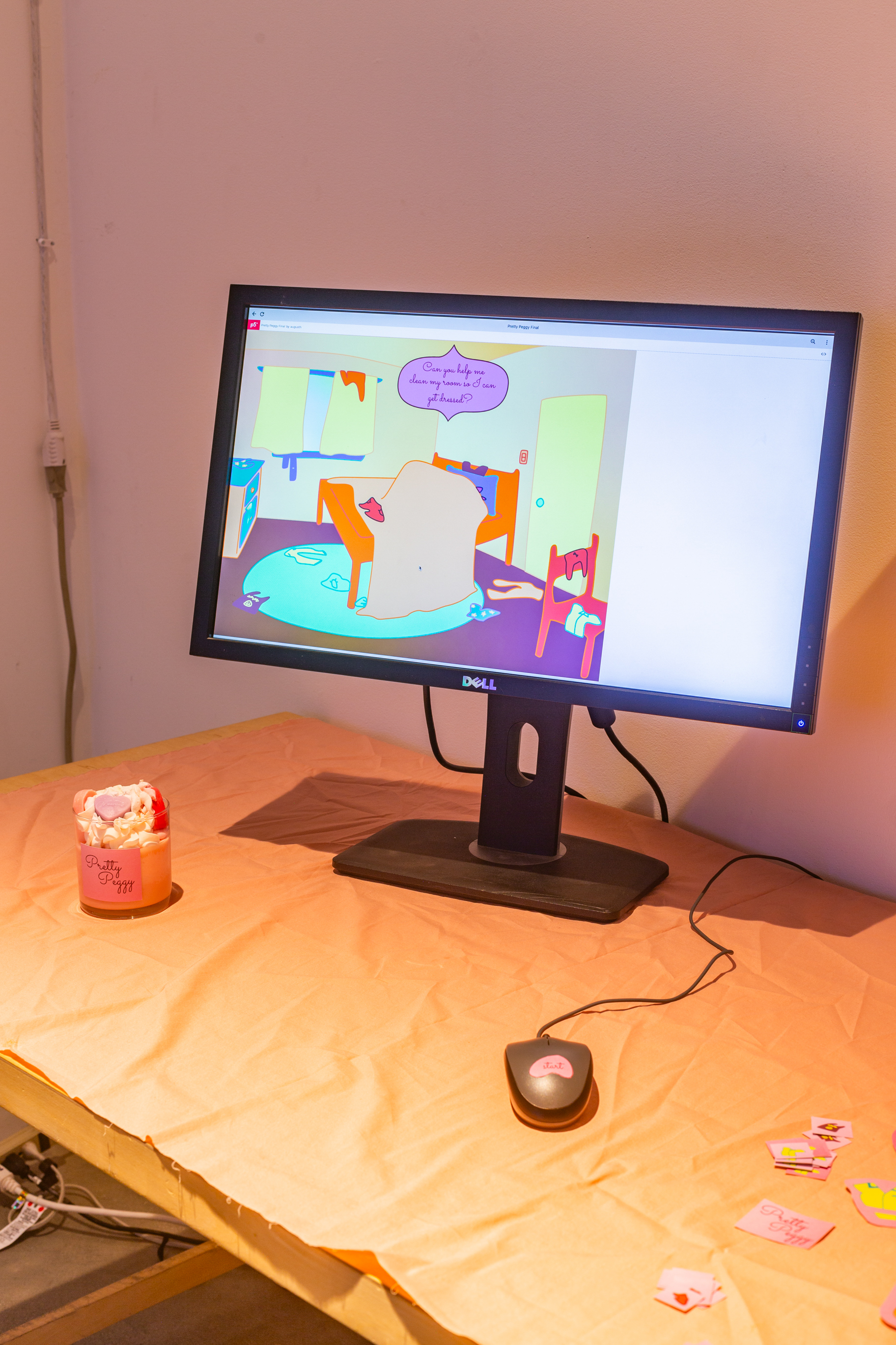 A table in a white space with a computer, a mouse, and a candle with candy in it. The computer has an interactive art piece displayed.